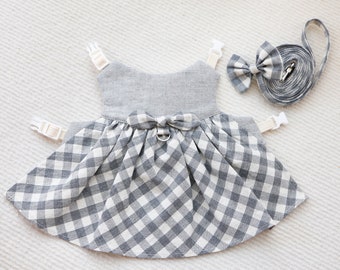 Puede incluir: Un vestido de perro de cuadros vichy gris y blanco con una pajarita y una correa a juego. El vestido tiene un diseño sencillo y sin mangas con una falda fruncida. La correa está hecha de un tejido suave y duradero.