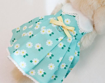 Puede incluir: Un vestido azul claro con ribete de encaje blanco y un lazo amarillo. El vestido tiene un estampado de margaritas y está diseñado para un perro pequeño.