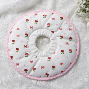 Puede incluir: Un collar de recuperaci&oacute;n para mascotas blanco con un estampado de cerezas y lazos. El collar tiene un ribete rosa a cuadros y un dise&ntilde;o suave y acolchado. La abertura central es para el cuello de la mascota.
