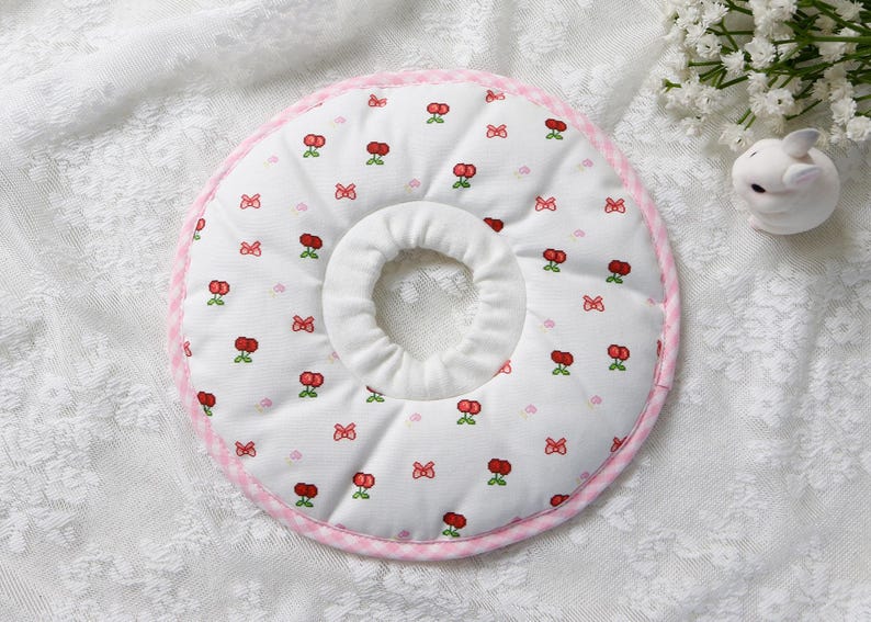 Puede incluir: Un collar de recuperaci&oacute;n para mascotas blanco con un estampado de cerezas y lazos. El collar tiene un ribete rosa a cuadros y un dise&ntilde;o suave y acolchado. La abertura central es para el cuello de la mascota.