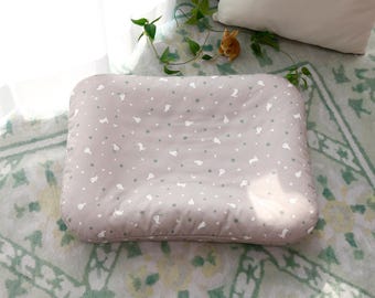 May include: A rectangular, light gray pillow with a pattern of white animal shapes and small gray dots. The pillow is on a patterned blanket with green and white floral designs. A small toy bunny and a white pillow are in the background.