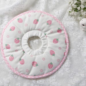 Puede incluir: Un collar de recuperaci&oacute;n para mascotas blanco con un estampado de fresas. El collar presenta fresas rosas y peque&ntilde;as flores sobre un fondo blanco. El borde est&aacute; adornado con tela de cuadros rosa.