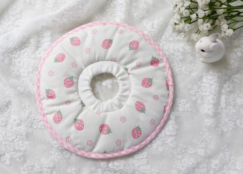 Puede incluir: Un collar de recuperaci&oacute;n para mascotas blanco con un estampado de fresas. El collar presenta fresas rosas y peque&ntilde;as flores sobre un fondo blanco. El borde est&aacute; adornado con tela de cuadros rosa.