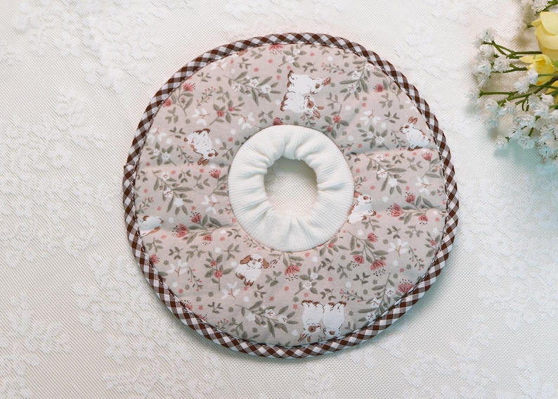 Puede incluir: Un collar de recuperaci&oacute;n para mascotas beige con un estampado floral y de conejitos. El collar tiene un centro blanco y un borde a cuadros marr&oacute;n y blanco. La tela es suave y acolchada.