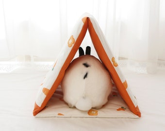 Peut inclure: Un lapin blanc avec des marques noires est assis dans un petit lit pour animal de compagnie en forme de tente orange et blanche. Le lit a une base blanche et des panneaux en tissu orange avec un motif de carottes et de fleurs.
