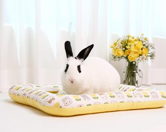 May include: A white rabbit with black ears and eyes rests on a yellow and white pet bed with a cartoon animal pattern. A small, yellow bowl is on the bed. A vase of yellow flowers sits in the background.