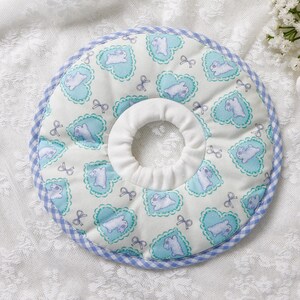 Puede incluir: Un collar de recuperaci&oacute;n acolchado para mascotas con un centro blanco y un exterior estampado en azul claro y blanco. El patr&oacute;n presenta corazones azul claro con un animal blanco en el interior y peque&ntilde;os lazos. El collar tiene un ribete de cuadros azul.