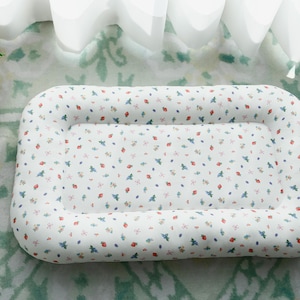 Könnte beinhalten: Ein rechteckiges Haustierbett mit weißem Hintergrund und einem Muster aus kleinen roten, blauen und grünen Blumenmustern. Das Bett hat einen gepolsterten, erhöhten Rand und liegt auf einem gemusterten Teppich.