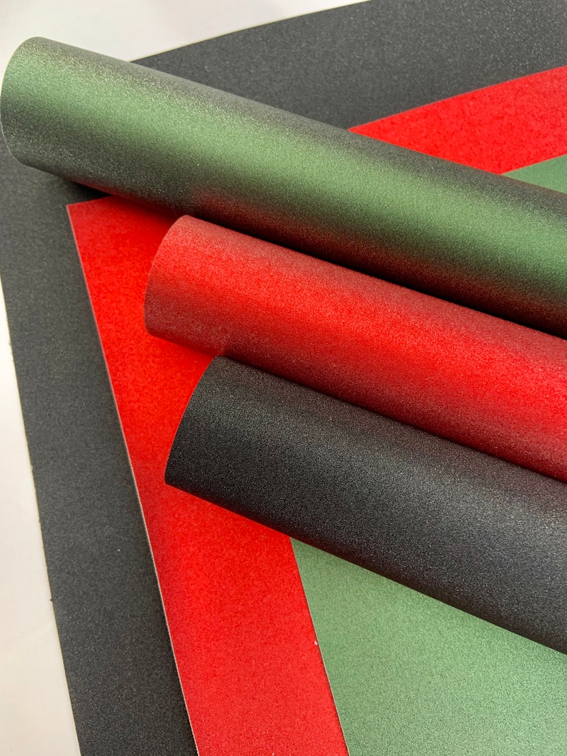 Op de afbeelding: Close-up van getextureerde papierrollen in groen, rood en zwart. De rollen zijn diagonaal gerangschikt, met de uiteinden zichtbaar. Het papier heeft een licht glinsterend uiterlijk. De achtergrond is effen wit.