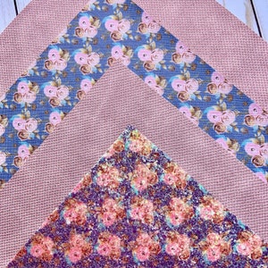 May include: An overhead shot displays three pieces of fabric. A textured, mauve-colored fabric forms the base. A blue fabric with pink floral patterns and a glittery, purple fabric with pink floral patterns are layered on top.