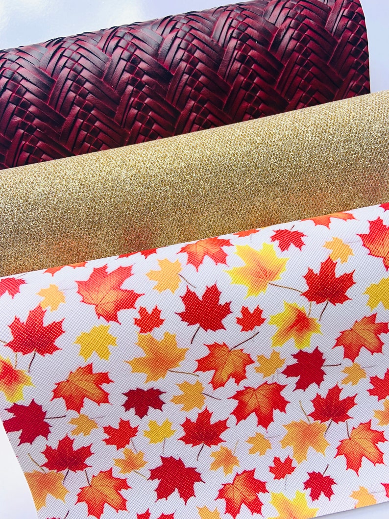 May include: Three rolls of fabric with different patterns. The top roll is a dark red woven fabric. The middle roll is a gold glitter fabric. The bottom roll is a white fabric with a red and yellow maple leaf pattern.