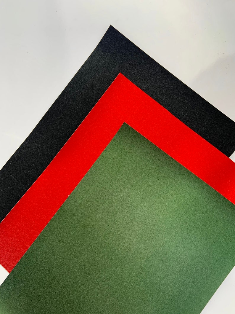 Op de afbeelding: Een diagonale compositie van drie vellen getextureerd papier in zwart, rood en olijfgroen. Het zwarte vel ligt bovenop, gevolgd door rood en groen, wat een visueel contrast cre&euml;ert. De textuur is goed zichtbaar.