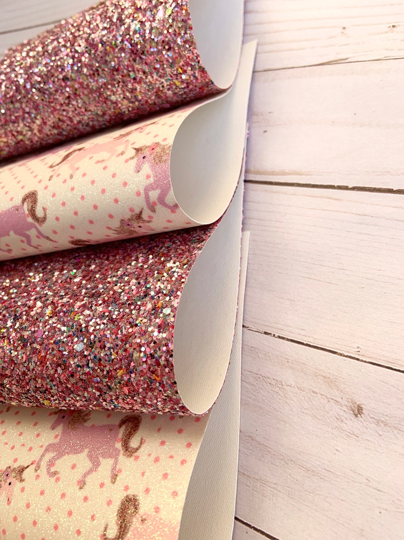 May include: Three sheets of pink glitter fabric with a white unicorn print. The fabric is folded in a stack.