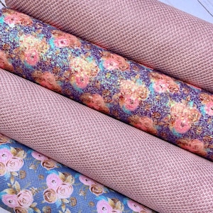 May include: Close-up of several rolls of fabric. Two rolls are a solid, textured, light pink. The other two rolls have floral patterns in pink, blue, and gold. The fabric appears to be faux leather or vinyl.