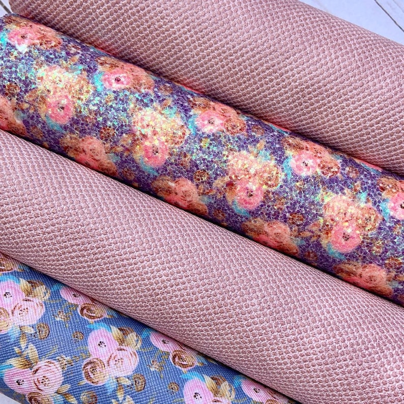 May include: Close-up of several rolls of fabric. Two rolls are a solid, textured, light pink. The other two rolls have floral patterns in pink, blue, and gold. The fabric appears to be faux leather or vinyl.