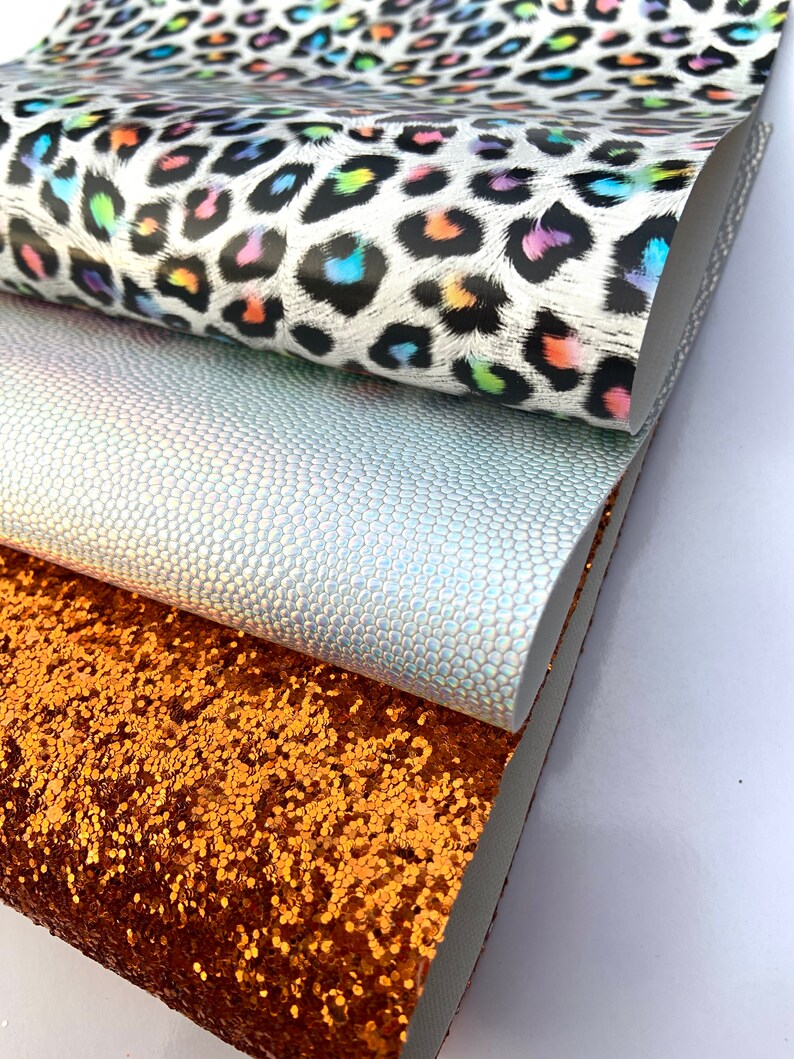 May include: Three different fabric swatches stacked on top of each other. The top swatch is a white fabric with a black and rainbow leopard print. The middle swatch is a light gray fabric with a subtle iridescent texture. The bottom swatch is a copper colored fabric with a glitter finish.