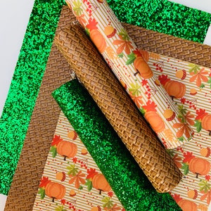 May include: An assortment of craft materials featuring a green glitter sheet, a brown textured sheet, and a rolled sheet with a pumpkin and leaf pattern. The materials are suitable for fall-themed crafts.