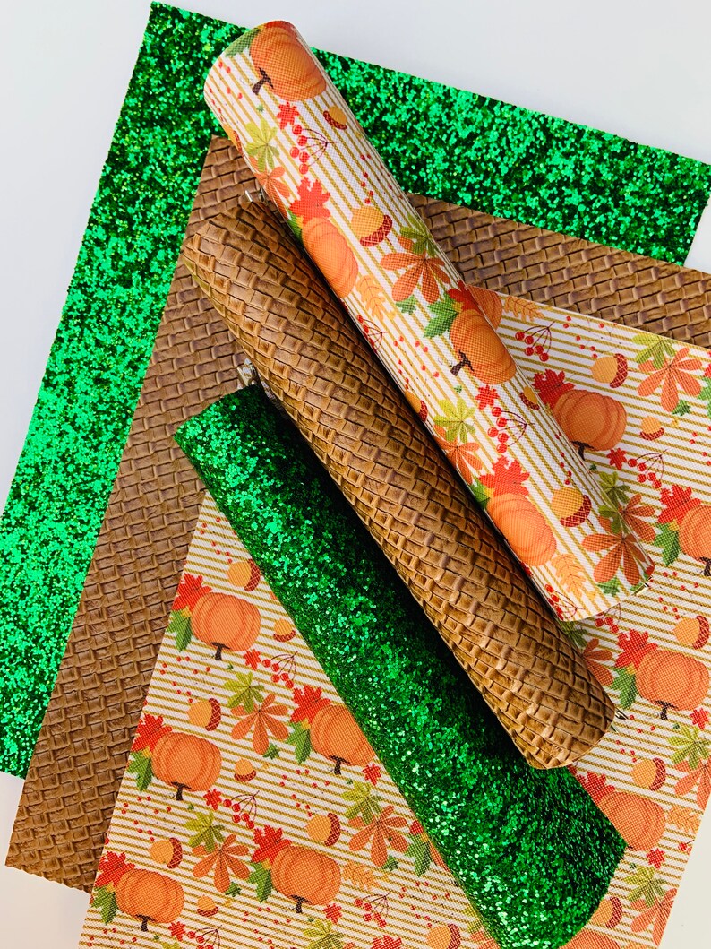 May include: An assortment of craft materials featuring a green glitter sheet, a brown textured sheet, and a rolled sheet with a pumpkin and leaf pattern. The materials are suitable for fall-themed crafts.