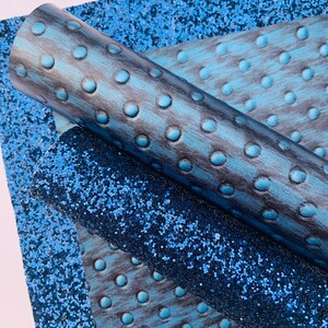 Op de afbeelding: Twee rollen blauwe kunstleer stof. E&eacute;n rol heeft een textuur, gegraveerd patroon van kleine cirkels. De andere rol heeft een glinsterende, glitter afwerking.