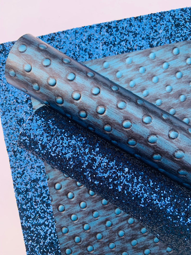 Op de afbeelding: Twee rollen blauwe kunstleer stof. E&eacute;n rol heeft een textuur, gegraveerd patroon van kleine cirkels. De andere rol heeft een glinsterende, glitter afwerking.