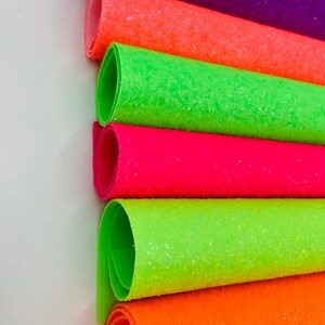 Pu&ograve; includere: Sei rotoli di tessuto glitterato in vari colori neon, tra cui verde, arancione, rosa e viola. Il tessuto &egrave; arrotolato e impilato uno sopra l'altro.