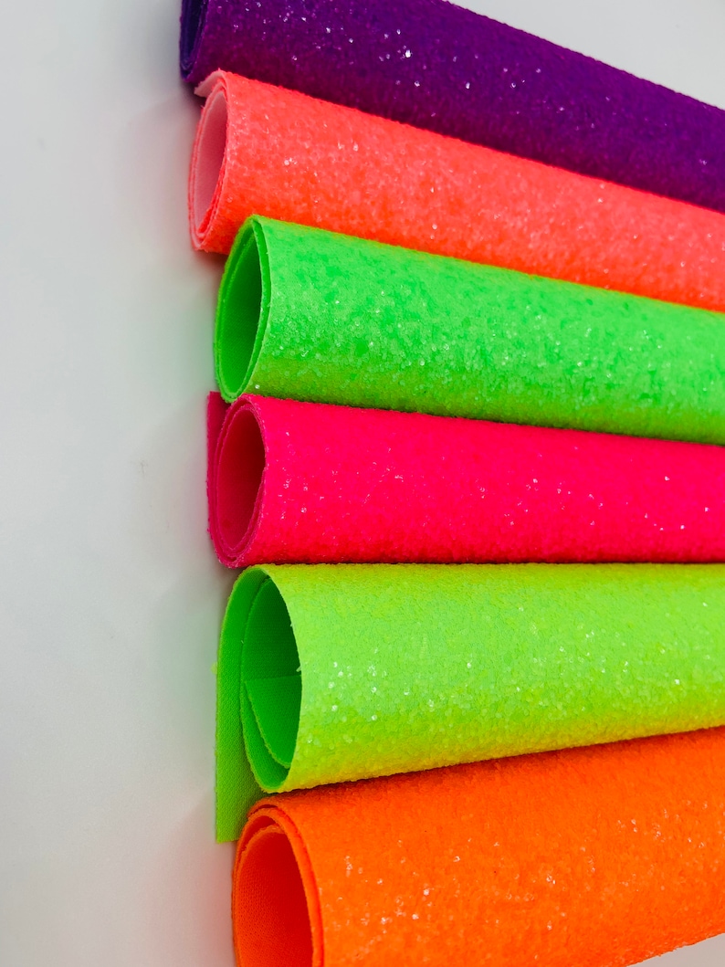 Pu&ograve; includere: Sei rotoli di tessuto glitterato in vari colori neon, tra cui verde, arancione, rosa e viola. Il tessuto &egrave; arrotolato e impilato uno sopra l'altro.