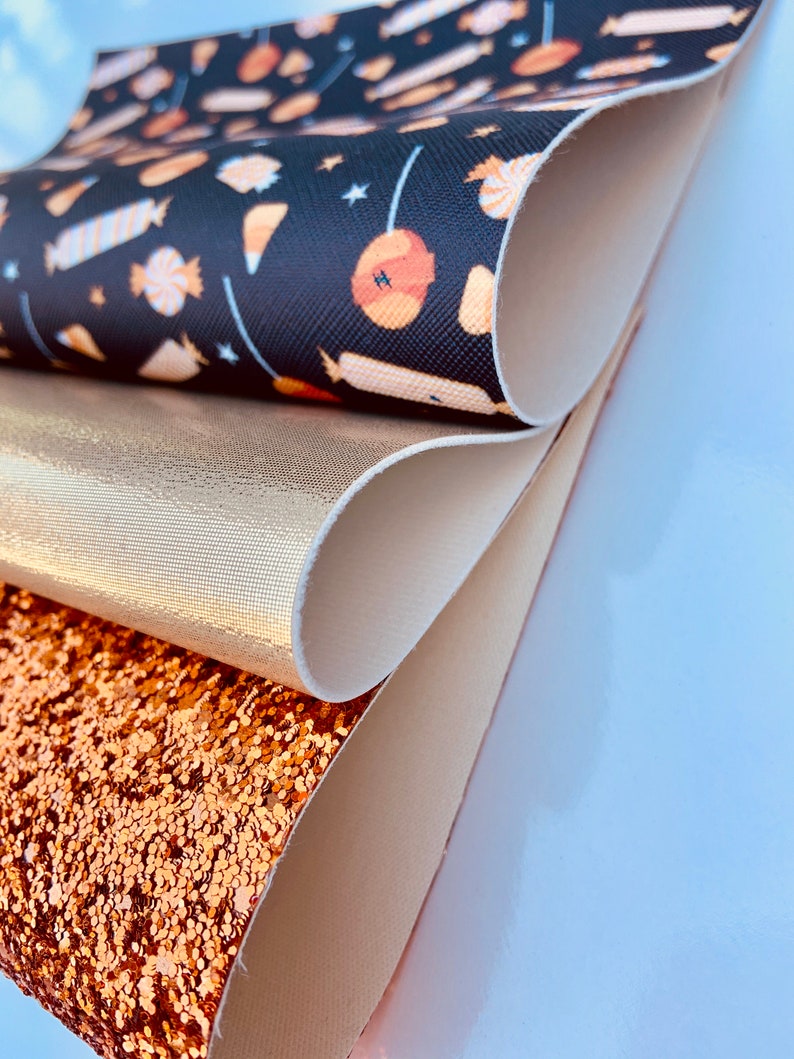 May include: A stack of four fabric swatches. The top swatch is black with a white and orange candy pattern. The second swatch is a metallic gold. The third swatch is a light beige. The bottom swatch is a sparkly copper.