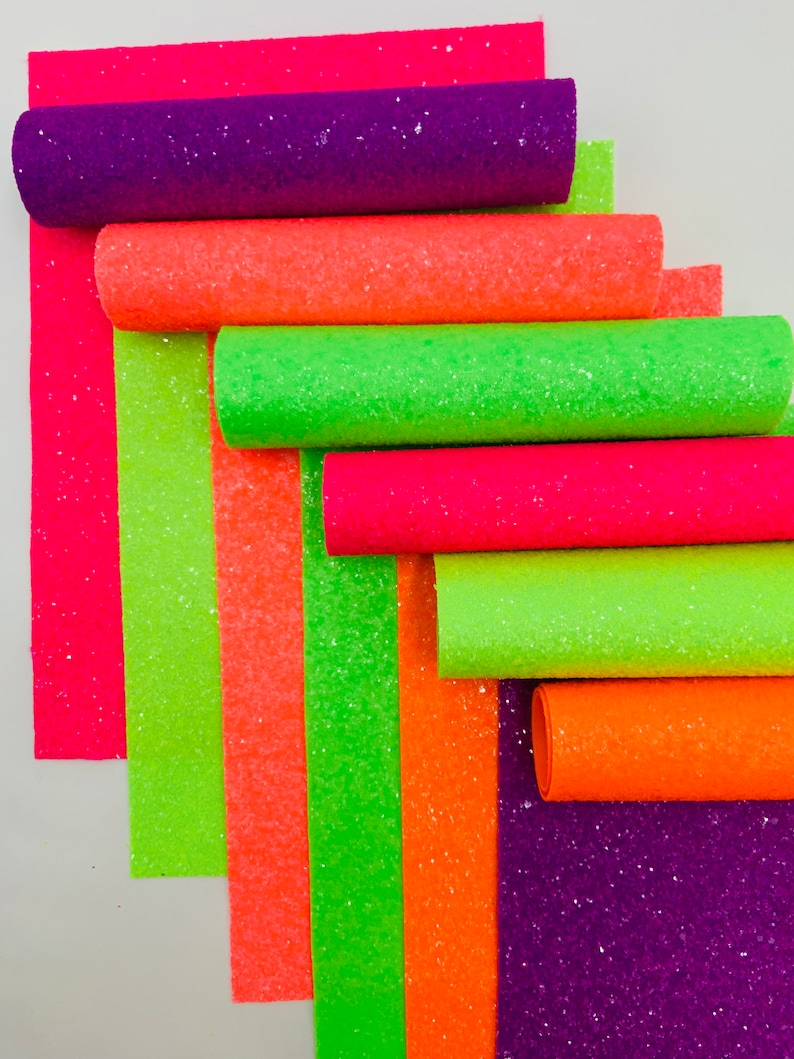 Pu&ograve; includere: Un rotolo di tessuto glitterato in vari colori neon, tra cui rosa, arancione, verde e viola. Il tessuto &egrave; arrotolato e le estremit&agrave; sono visibili.
