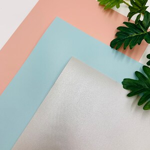 May include: Three sheets of faux leather in different colors: light pink, light blue, and silver. The sheets are arranged in a triangular shape with the silver sheet at the bottom.