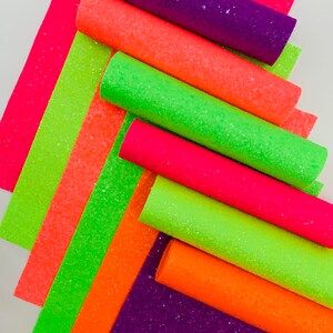 Pu&ograve; includere: Una collezione di rotoli di tessuto glitterato verde neon, arancione e rosa. I rotoli sono impilati uno sopra l'altro, con i rotoli verdi in basso e i rotoli rosa in alto.