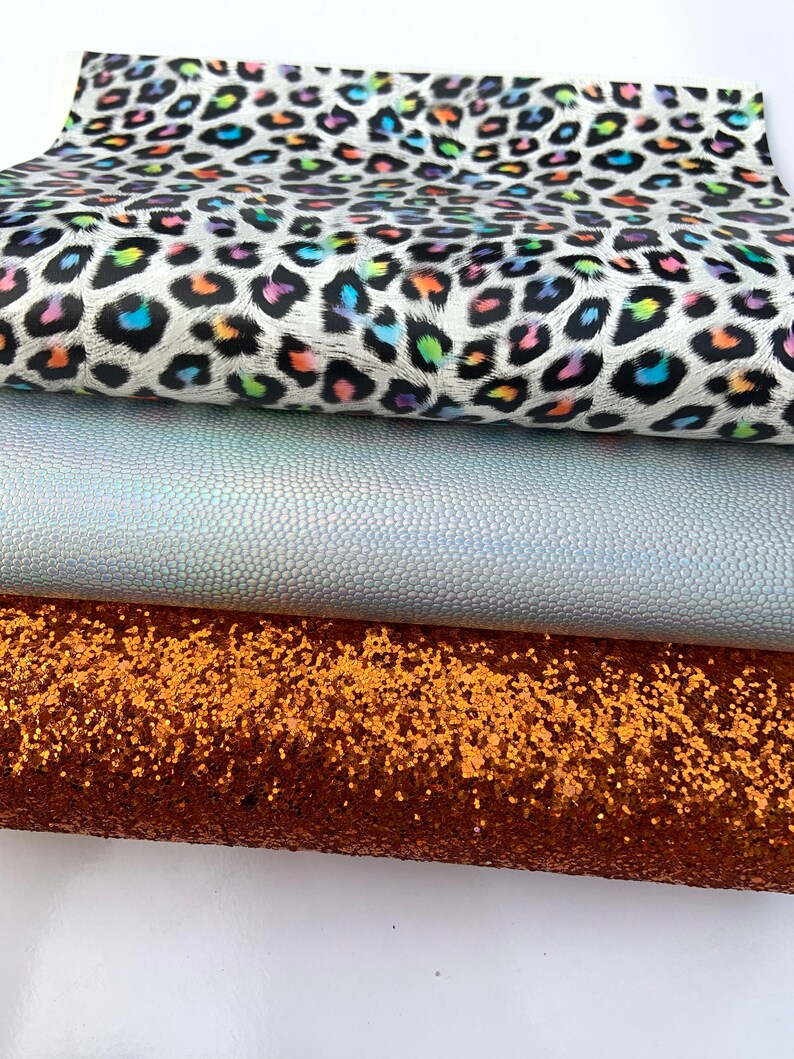 May include: Three fabric swatches: a white fabric with a rainbow leopard print, a light blue iridescent fabric with a textured surface, and a copper glitter fabric.