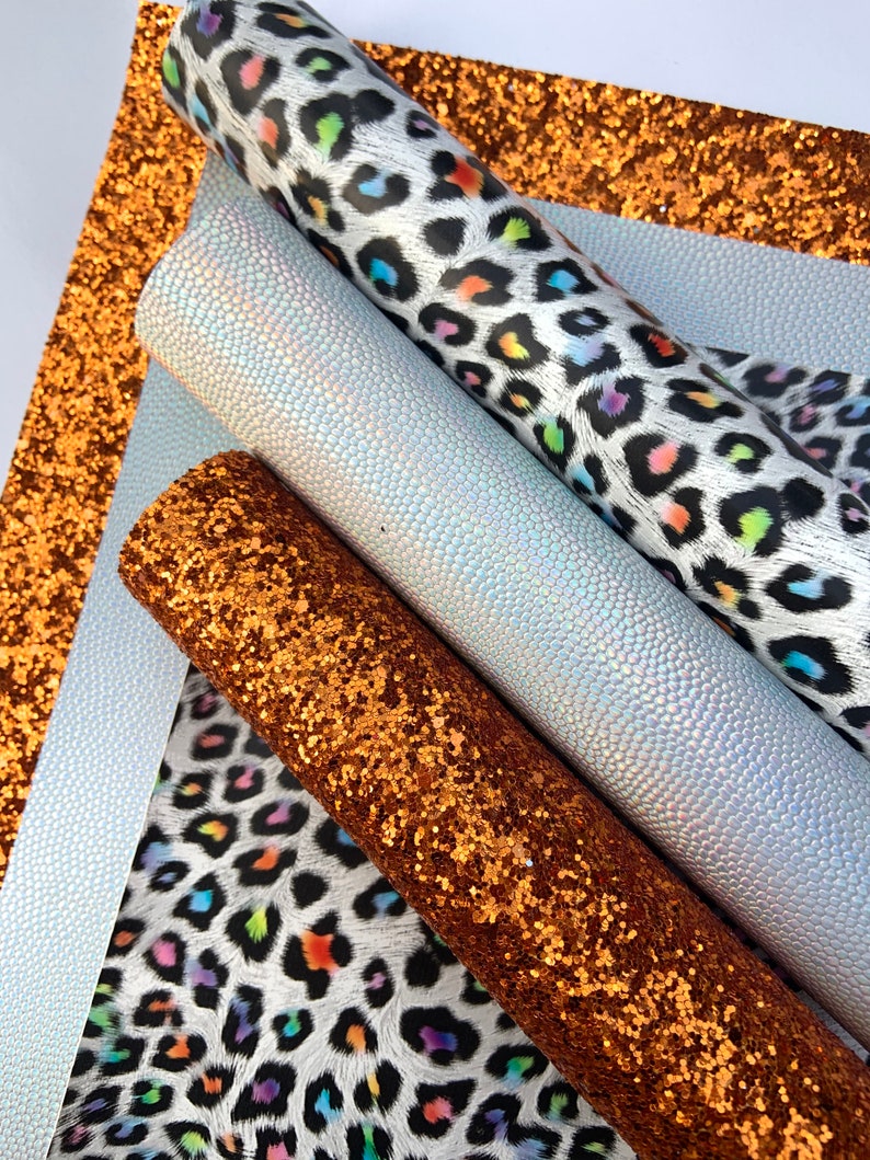 May include: Four sheets of faux leather in different patterns and colors. One sheet is a white and black leopard print with rainbow colored spots. Another sheet is a iridescent white with a textured surface. A third sheet is a copper glitter fabric. The fourth sheet is a solid copper color.