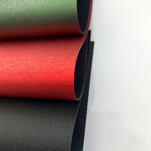 Op de afbeelding: Close-up van drie vellen textiel in groen, rood en zwart. De vellen zijn gelaagd, waardoor een glimp van de zwarte onderkant te zien is. Het materiaal heeft een licht reflecterend, gespikkeld oppervlak.
