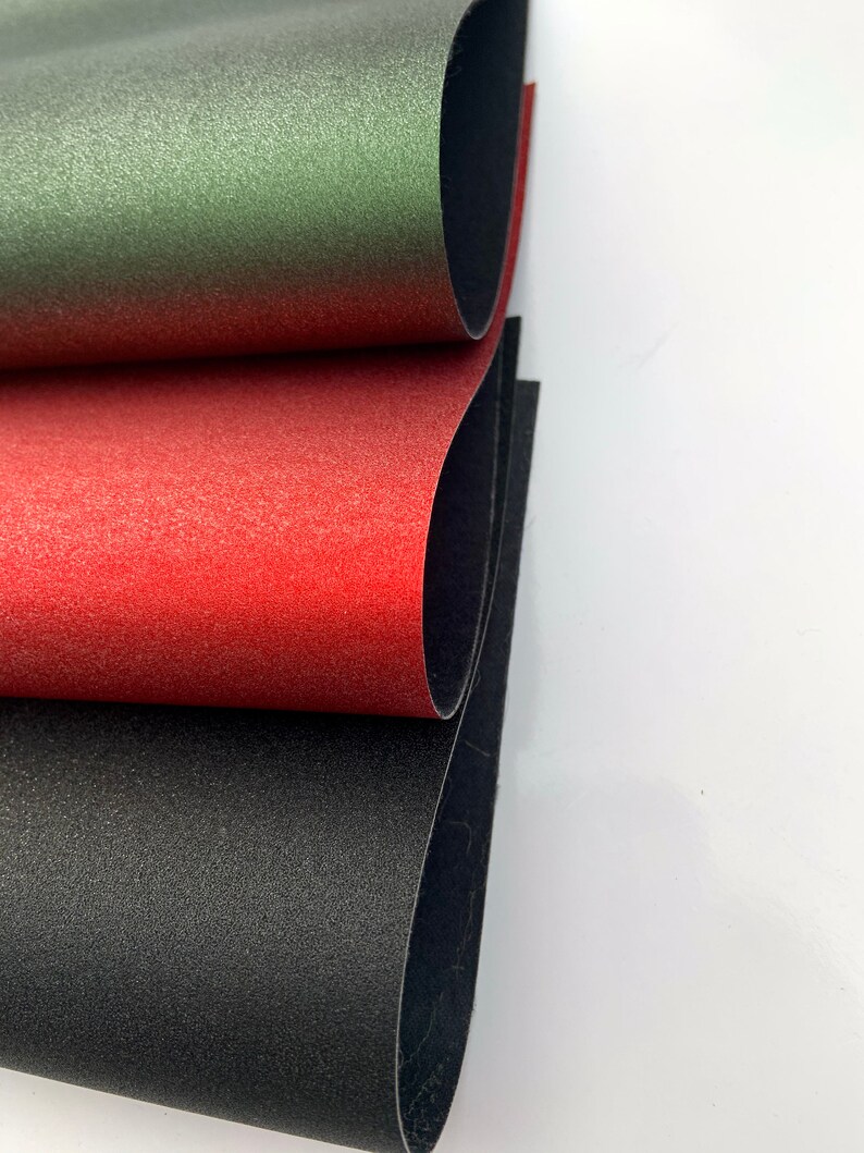 Op de afbeelding: Close-up van drie vellen textiel in groen, rood en zwart. De vellen zijn gelaagd, waardoor een glimp van de zwarte onderkant te zien is. Het materiaal heeft een licht reflecterend, gespikkeld oppervlak.