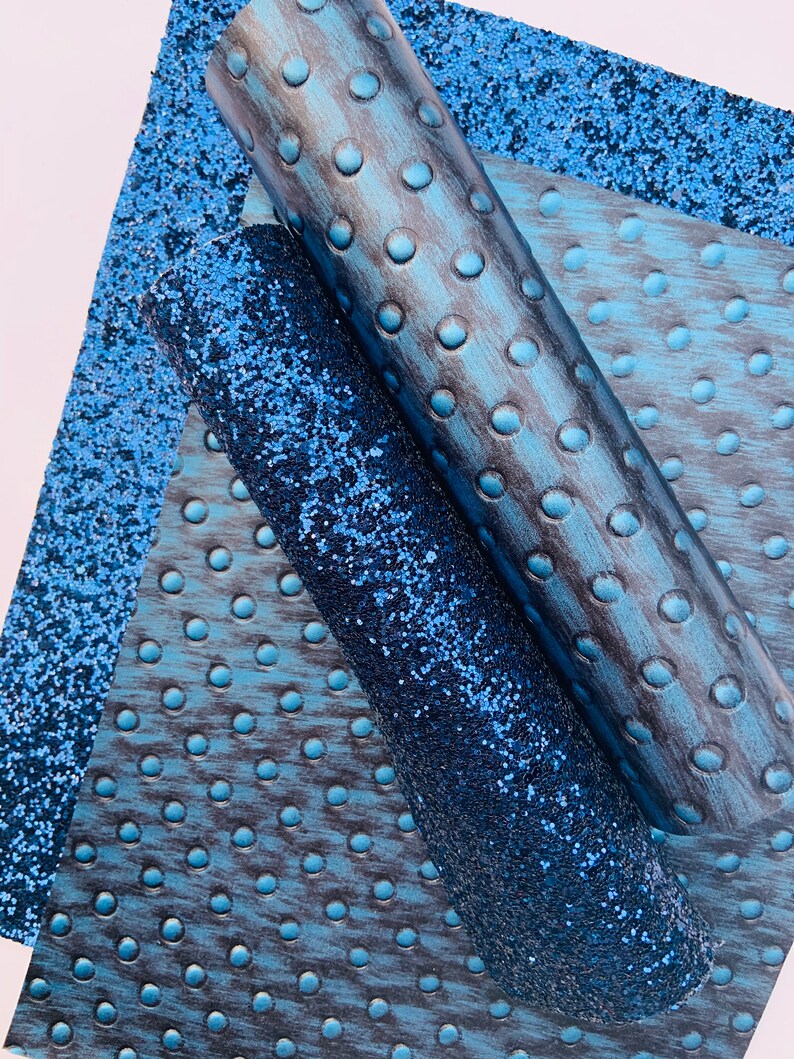 Op de afbeelding: Drie vellen blauw kunstleer met verschillende texturen. Een vel is een effen blauwe glitter, een ander is een blauw en zwart houtnerf met een stippenpatroon, en de derde is een blauw en zwart houtnerf met een groter stippenpatroon.