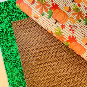 May include: An assortment of craft materials featuring a green glitter sheet, a brown woven texture, and a fall-themed pattern. The fall pattern includes orange pumpkins, autumn leaves, and acorns on a striped background.