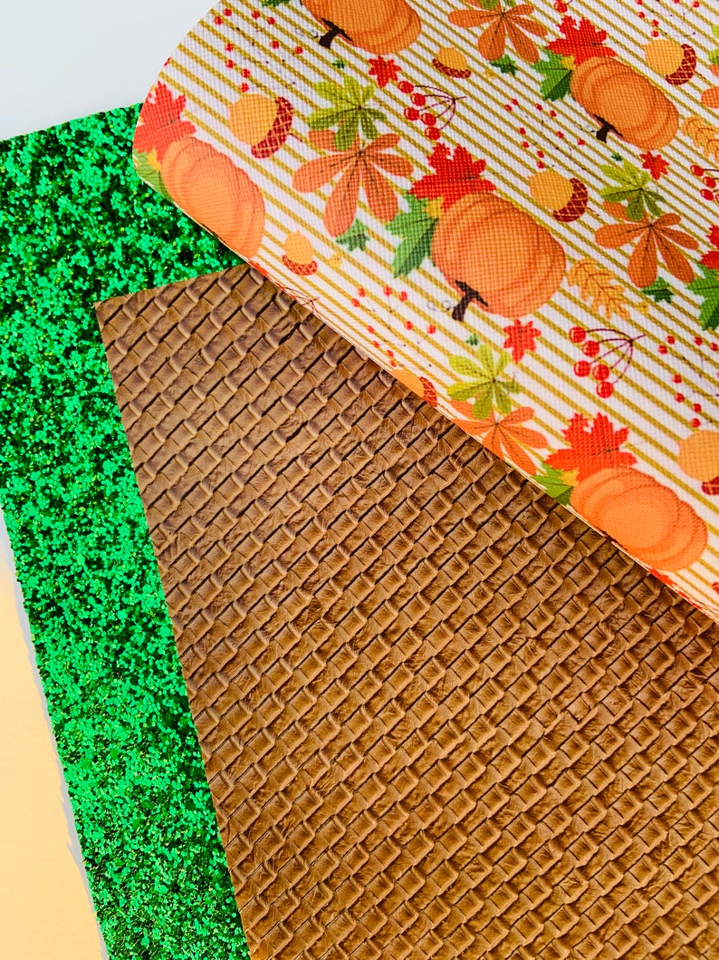May include: An assortment of craft materials featuring a green glitter sheet, a brown woven texture, and a fall-themed pattern. The fall pattern includes orange pumpkins, autumn leaves, and acorns on a striped background.