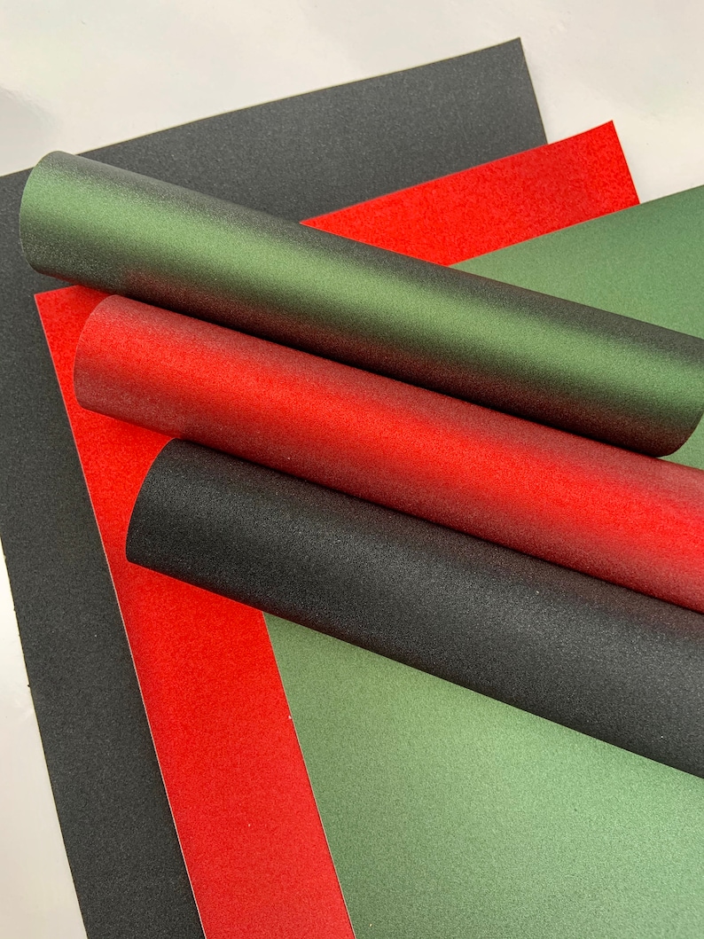 Op de afbeelding: Een verzameling getextureerd papier in rood, groen en zwart. Sommige vellen zijn opgerold, andere plat. Het papier heeft een subtiele glans, wat wijst op een metallic afwerking. Ideaal voor knutsel- en decoratieprojecten.