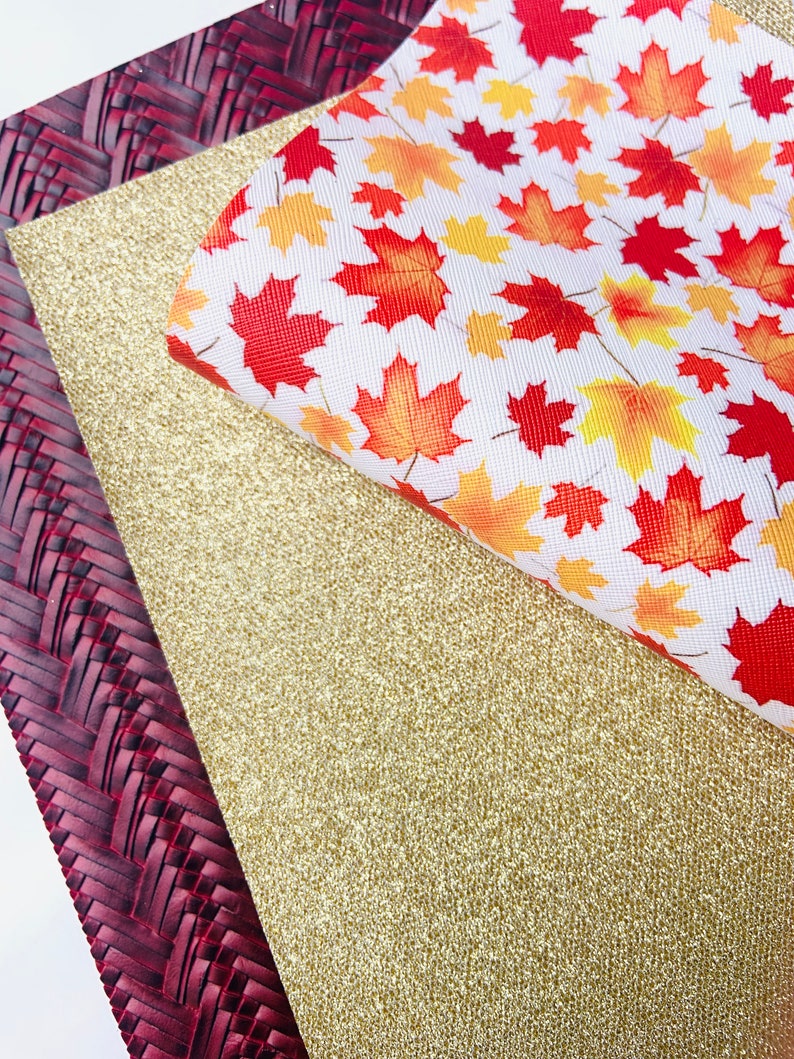 May include: Three sheets of fabric, one with a woven pattern in burgundy, one with a gold glitter finish, and one with a white background and a pattern of red, orange, and yellow maple leaves.