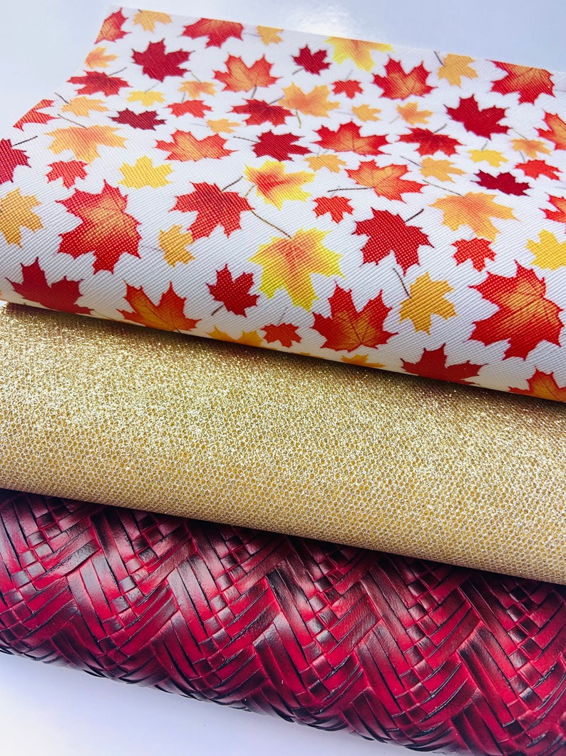 May include: Three pieces of fabric with different textures and patterns. The top piece is white with a red and yellow maple leaf pattern. The middle piece is gold glitter fabric. The bottom piece is a dark red woven fabric.