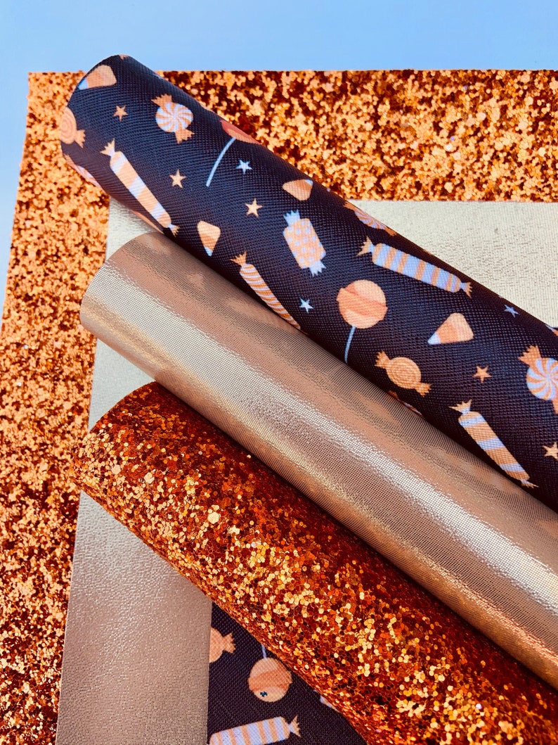 May include: A set of four craft supplies featuring a black fabric with a white and orange candy pattern, a gold glitter fabric, a copper glitter fabric, and a gold metallic fabric.