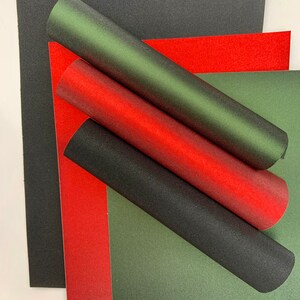 Op de afbeelding: Een assortiment getextureerd papier in rood, groen en zwart. Gerolde papieren buizen in bijpassende kleuren zijn over de platte vellen gerangschikt. Het papier heeft een subtiele glitterafwerking, wat een feestelijke en decoratieve uitstraling cre&euml;ert.