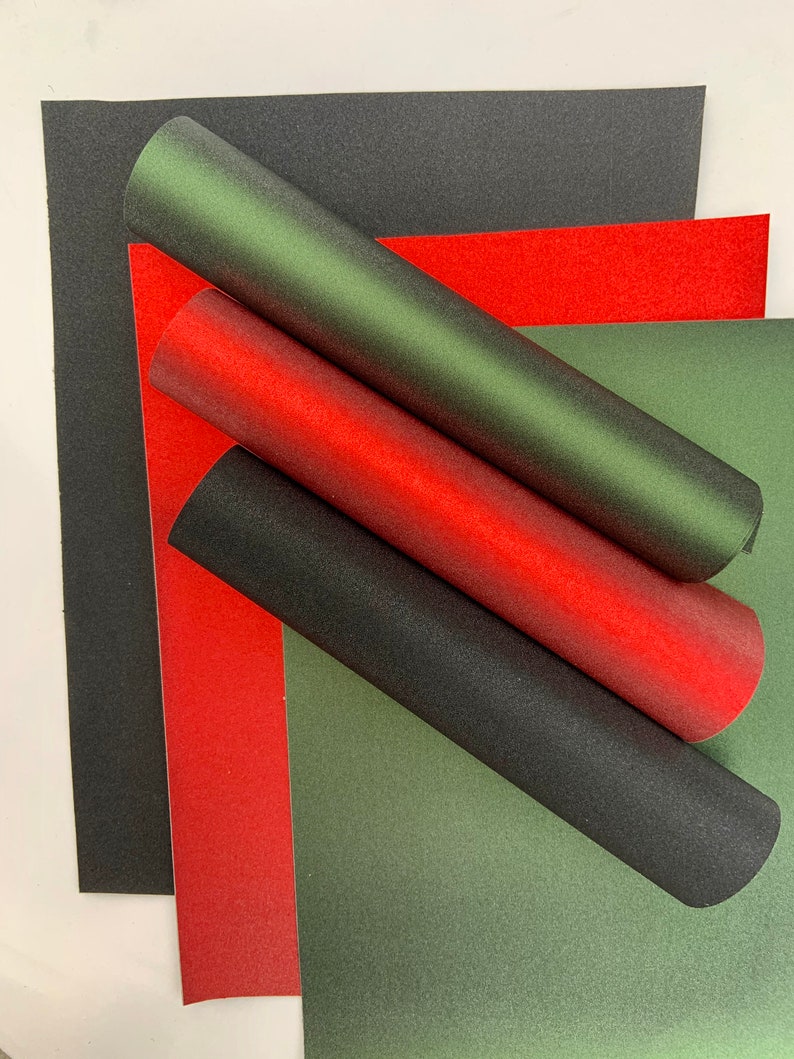 Op de afbeelding: Een assortiment getextureerd papier in rood, groen en zwart. Gerolde papieren buizen in bijpassende kleuren zijn over de platte vellen gerangschikt. Het papier heeft een subtiele glitterafwerking, wat een feestelijke en decoratieve uitstraling cre&euml;ert.