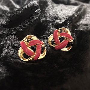 Puede incluir: Un par de pendientes de tono dorado con un diseño de nudo. Los nudos están acentuados con esmalte rosa.