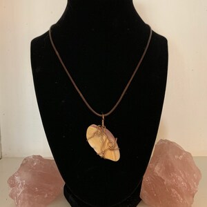 May include: A handcrafted pendant necklace featuring a light-colored stone wrapped in copper wire. The stone is suspended from a brown cord necklace. The necklace is displayed on a black mannequin. Two pink quartz crystals are on either side.