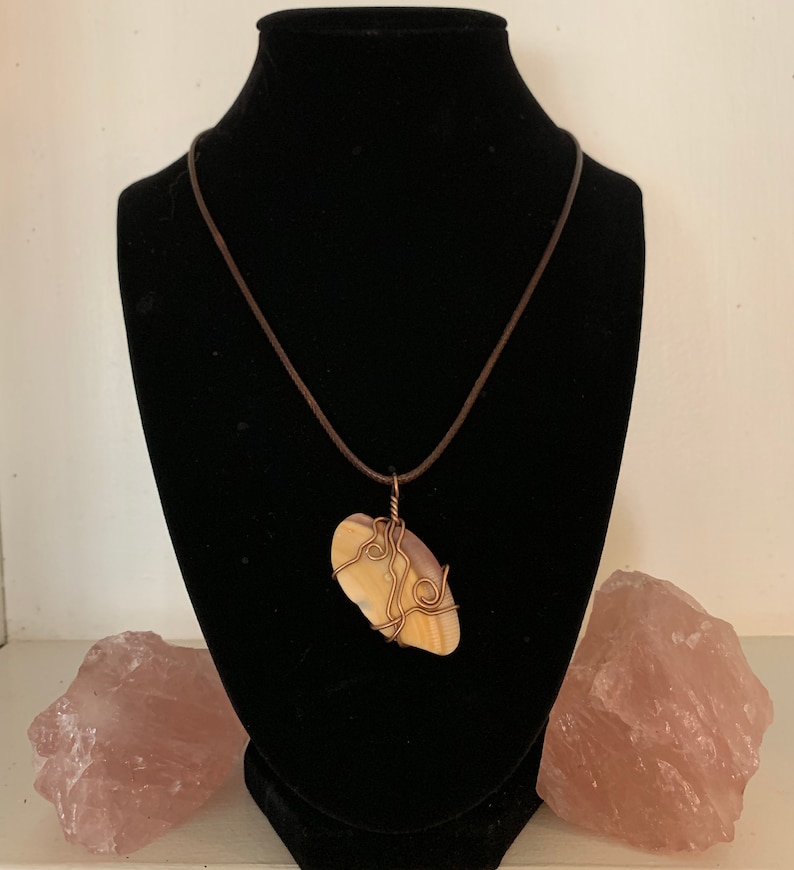 May include: A handcrafted pendant necklace featuring a light-colored stone wrapped in copper wire. The stone is suspended from a brown cord necklace. The necklace is displayed on a black mannequin. Two pink quartz crystals are on either side.