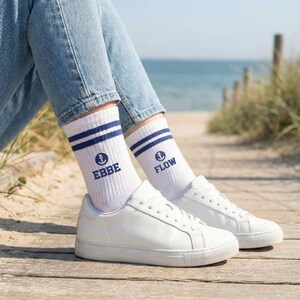 Könnte beinhalten: Weiße Sportsocken mit marineblauen Streifen und den Wörtern "EBBE" und "FLOW" in Marineblau sowie einem Ankersymbol. Die Socken werden mit weißen Turnschuhen und hellblauen Jeans getragen. Der Hintergrund zeigt einen Strand und einen Holzsteg.