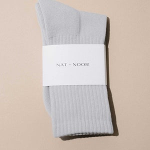 May include: A pair of gray ribbed socks folded and packaged with a white label that reads "NAT + NOOR".