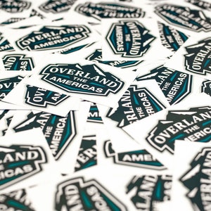 May include: A pile of white square stickers with a black and teal logo that says "Overland The Americas".