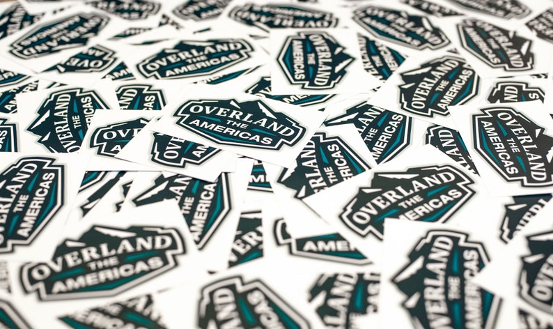 May include: A pile of white square stickers with a black and teal logo that says "Overland The Americas".